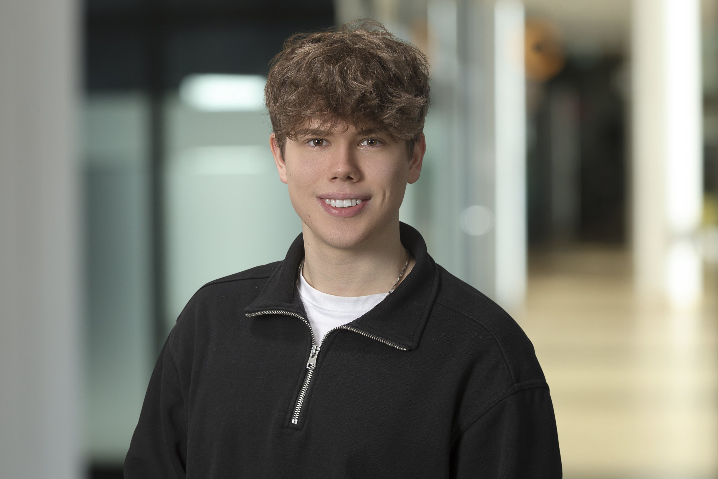 Portrait junger Mann mit kurzen welligem Haar und schwarzem Pullover mit Reißverschluss und weißem Shirt
